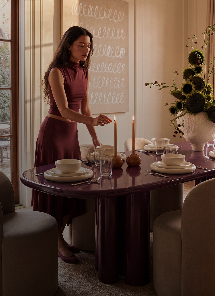 A woman in a matching burgundy two-piece outfit stands beside a glossy dark purple dining table, lighting a tall candle in a warmly lit interior setting.