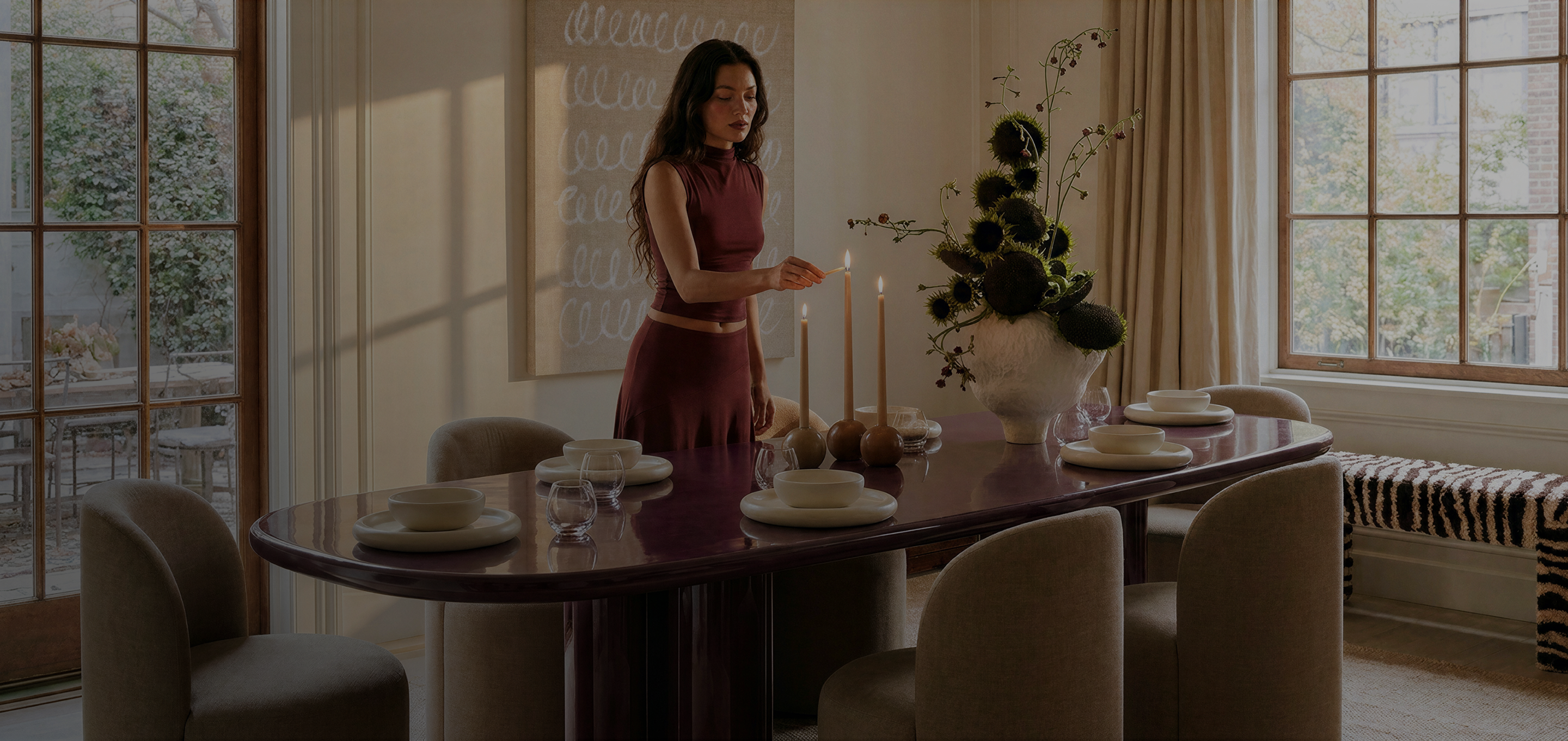 A woman in a sleeveless rust-brown dress stands at a dining table with lit taper candles, set plates, and a large floral arrangement in a sunlit dining room.