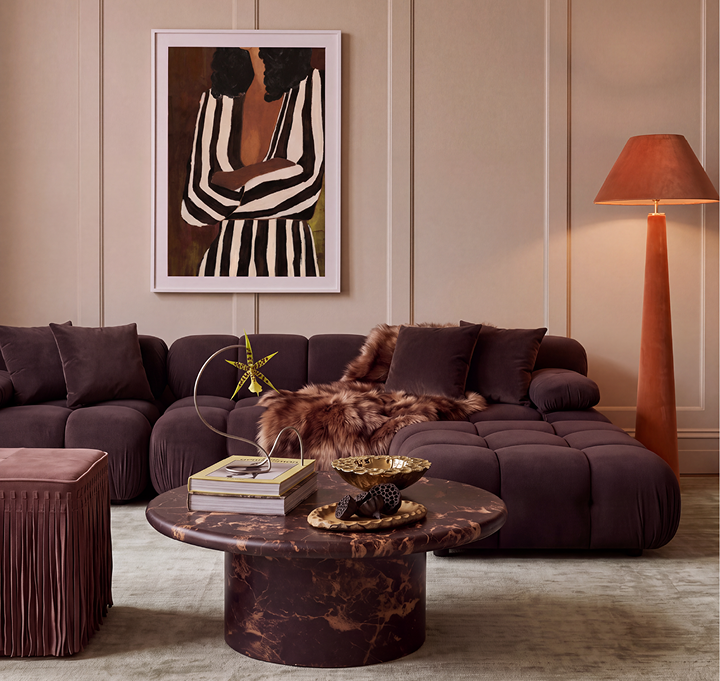 A styled living room featuring a deep purple modular sectional sofa with a brown faux-fur throw, a round dark marble coffee table, a matching pleated ottoman, and a tall floor lamp. A framed abstract art print hangs on the beige paneled wall above the sofa.